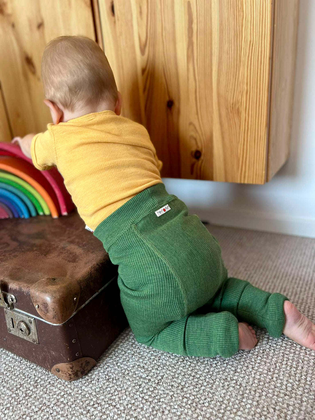 A baby wearing a yellow long-sleeve top and wool longies green ribbed pants crawls on a textured carpet next to a vintage brown suitcase, reaching toward a rainbow toy in front of a wooden cabinet.