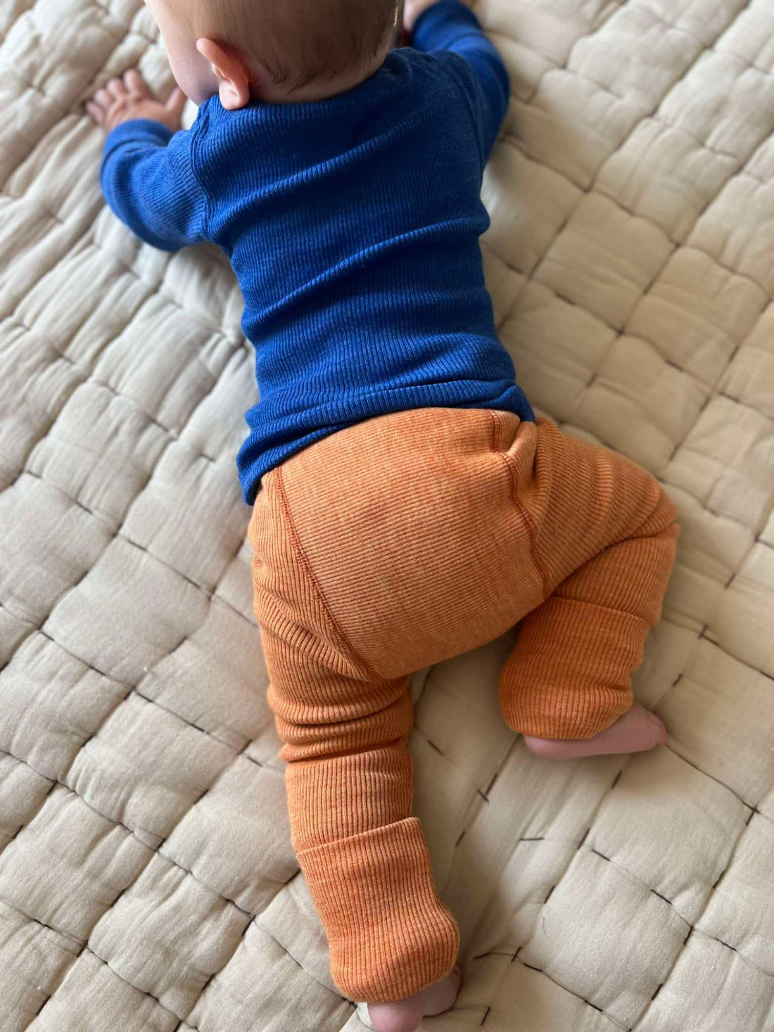 A baby wearing a yellow long-sleeve top and wool longies green ribbed pants crawls on a textured carpet next to a vintage brown suitcase, reaching toward a rainbow toy in front of a wooden cabinet.