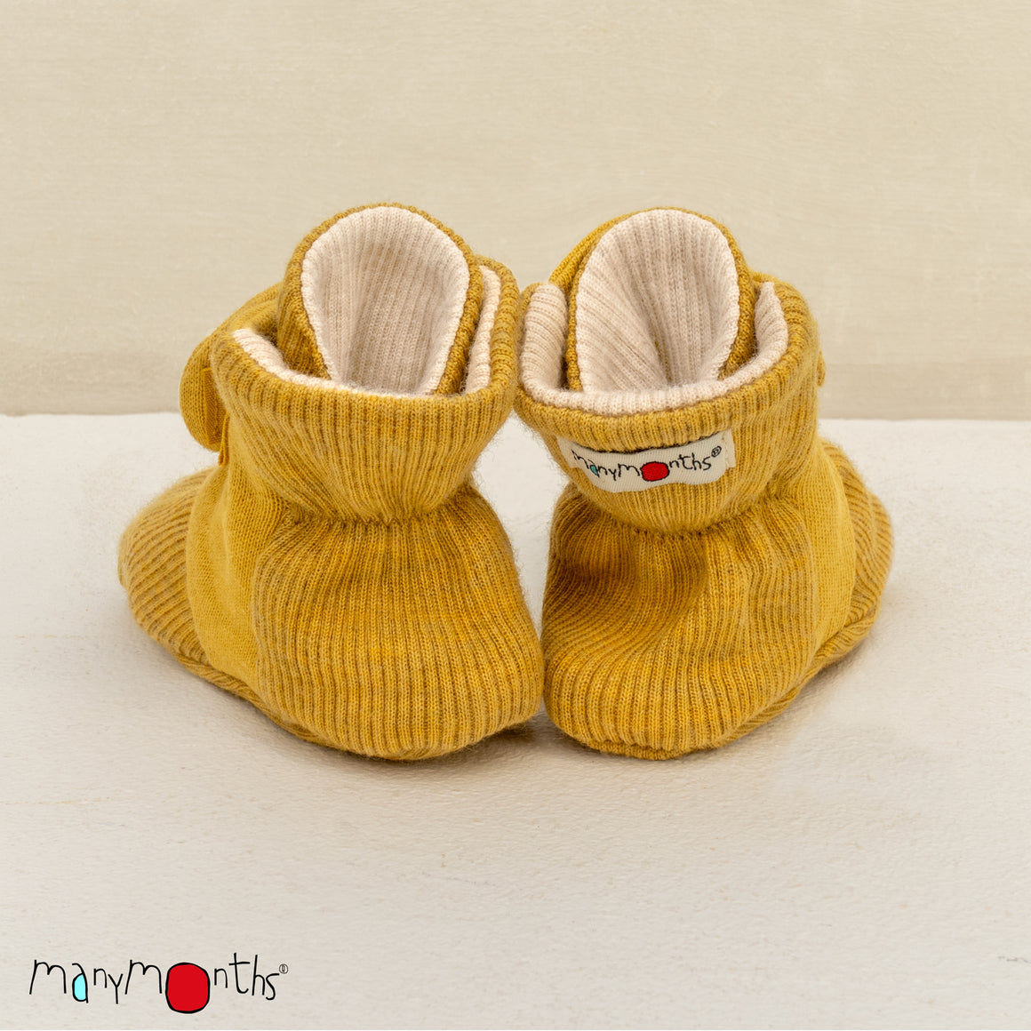 A pair of mustard yellow ribbed WOOL  knit baby booties with elasticized wrap-around straps, displayed on a white surface against a light beige wall. The Many Months brand logo is visible in the bottom right corner.