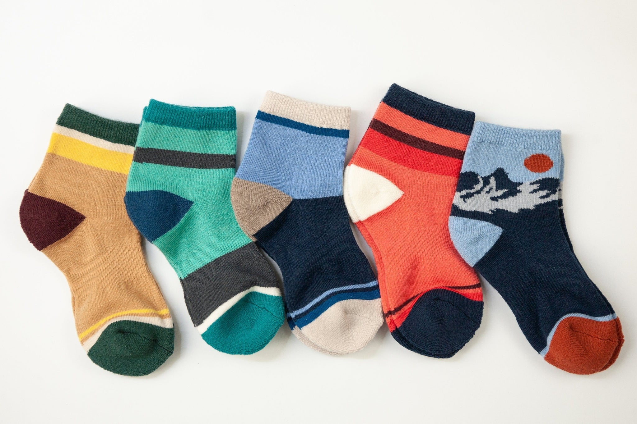 Six pairs of multi-colored merino wool crew socks arranged in a row on a plain white background. The socks feature various patterns and color combinations, including stripes, blocks, and a mountain-and-sun graphic. All kids sizes shown