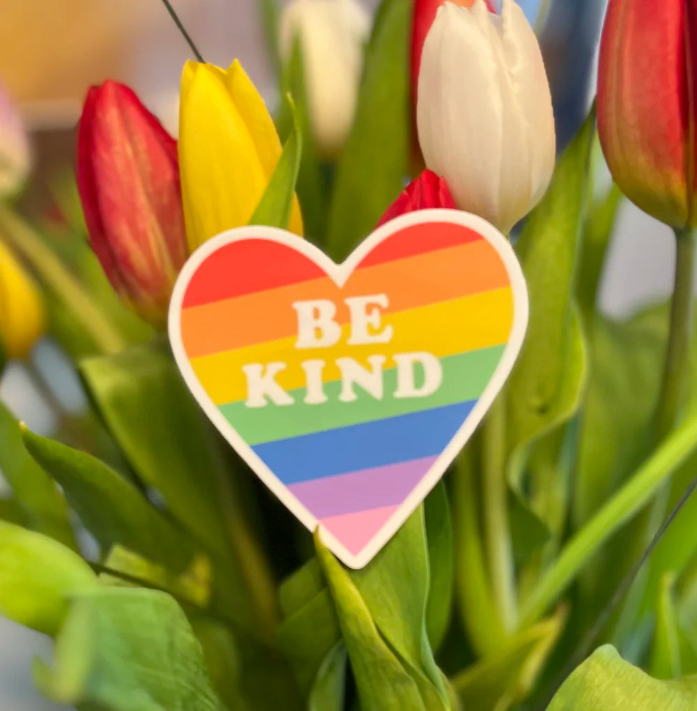 A bouquet of pink and red tulips in a clear glass vase, with a red heart-shaped card in the center that reads “BE KIND” in white letters.
